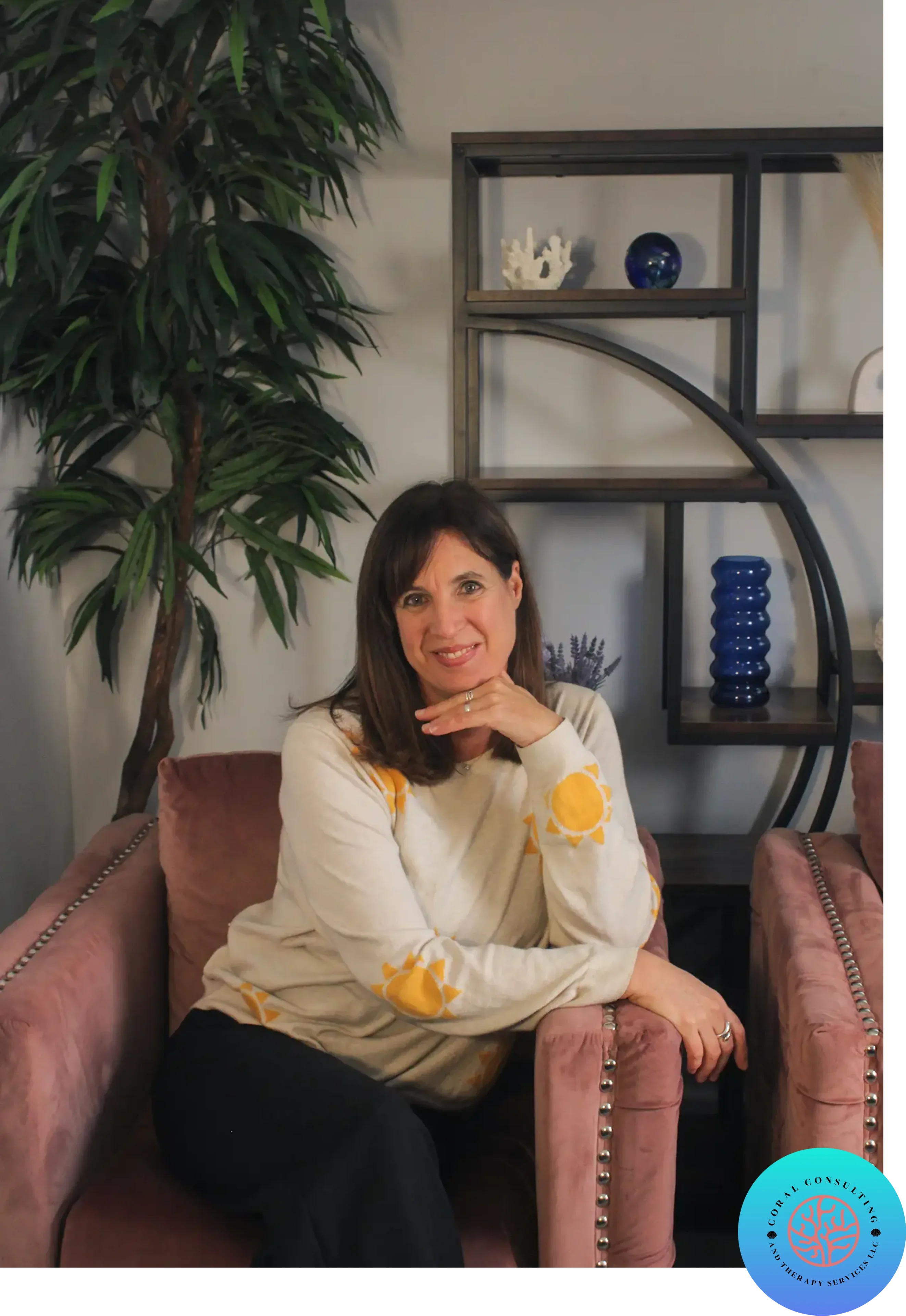 A woman sitting on a pink couch with a plant and shelves in the background.